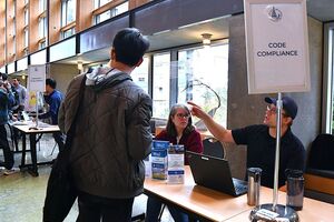 Two people sit at a table with a person standing facing them on the other side. One person is pointing. The sign on the table says Code Compliance.
