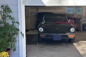 A vehicle parked in a garage. A blanket and tools are visible on the hood. Greenery is outside the garage door.